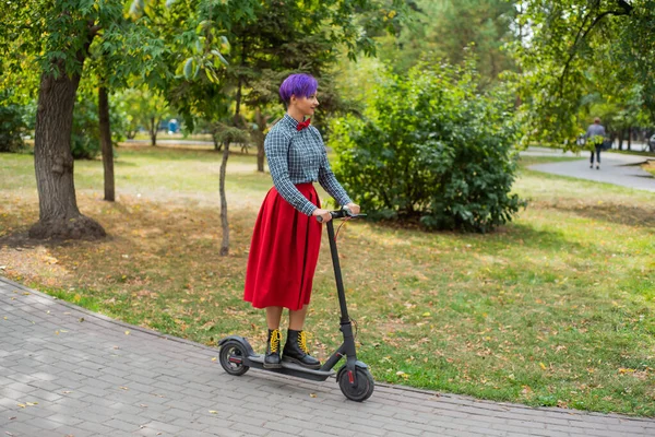 Egy fiatal nő, lila hajú lovagol egy elektromos robogó egy parkban. Stílusos lány egy kockás ing, egy hosszú vörös szoknya és egy csokornyakkendő a lovaglás a város körül egy modern eszköz. Csípő. — Stock Fotó