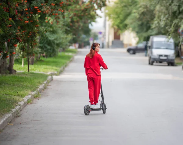 Rudowłosy dziewczyna w czerwonym dres napędza skuter elektryczny. Młoda kobieta w przewymiarowany ubrania jeździ po mieście i słucha muzyki za pomocą słuchawek bezprzewodowych. Widok z tyłu. — Zdjęcie stockowe