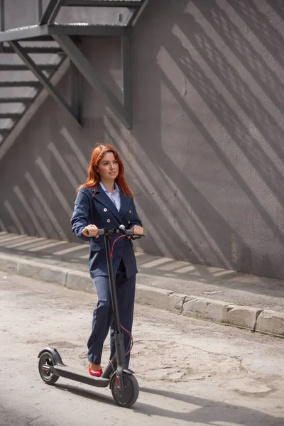 A red-haired girl in a jacket and trousers drives an electric scooter along the wall. A business woman in a pantsuit and red high heels rides around the city in a modern car. Office worker. Dress code