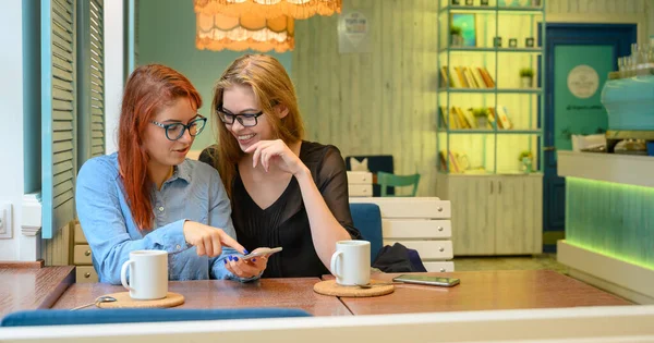 İki heyecanlı genç kız cep telefonu kullanıyor, bir kafede oturuyor ve parmakla işaret ediyor. Gözlüklü kızıl saçlı kadın akıllı telefon kız arkadaşının komik fotoğraflarını gösteriyor. Kahve içerler..
