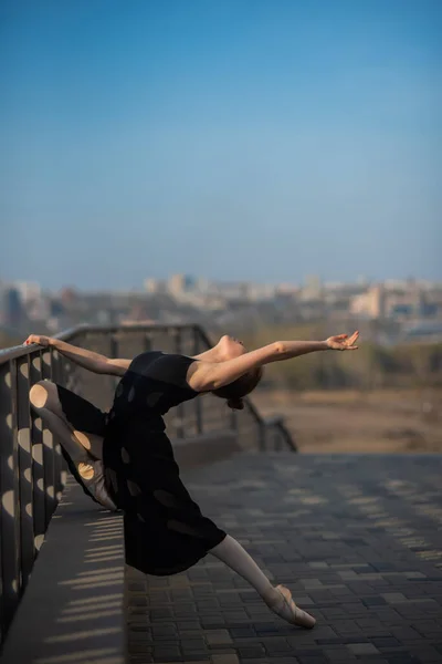 Ballerina in a tutu posing near the fence. Beautiful young woman in black dress and pointe dancing outside. Gorgeous ballerina demonstrates amazing stretching. — Stock Photo, Image