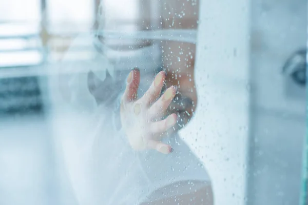 Mujer joven, posando detrás de vidrio transparente cubierto por gotas de agua. retrato femenino melancólico y triste. Rubia en una camiseta blanca toca el vidrio con gotas en el baño —  Fotos de Stock