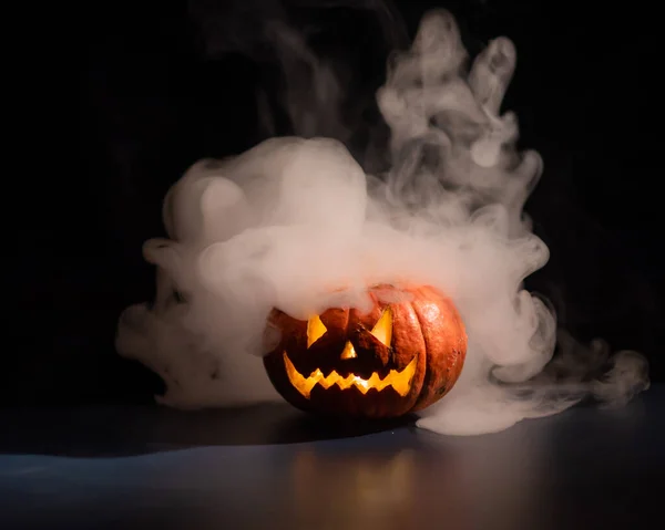 Halloween, apelsin pumpa med en skrämmande lysande ansikte på en mörk bakgrund. Tjock grå rök kommer ut och sprider sig över det svarta bordet. En närbild på en ficklampa. Dekorerat ljus stativ. — Stockfoto