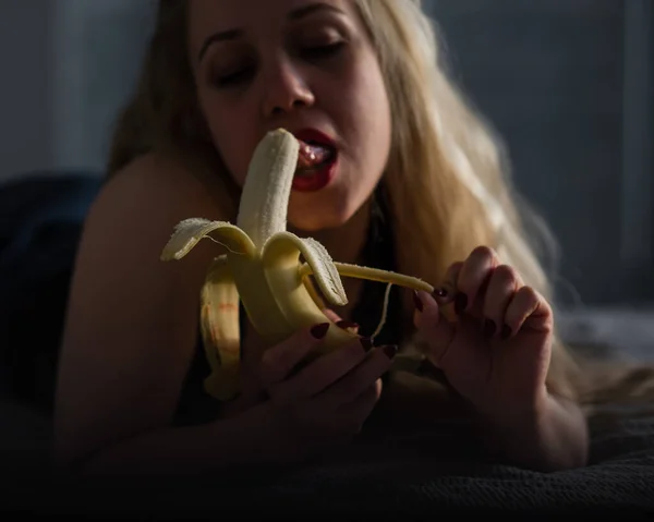 Ein Mädchen mit langen blonden Haaren schaut sexy auf eine große Banane. öffnet ihren Mund und präsentiert Oralsex — Stockfoto