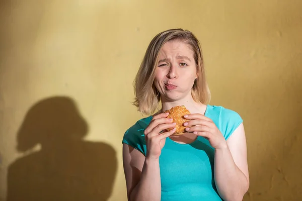 Portrét hezké dívky v zelených šatech venku a jíst Burger v teplém letním dnu. Krásná blondýnka si užívá rychlého občerstvení ve žluté zdi venku. Špatné stravní návyky. — Stock fotografie