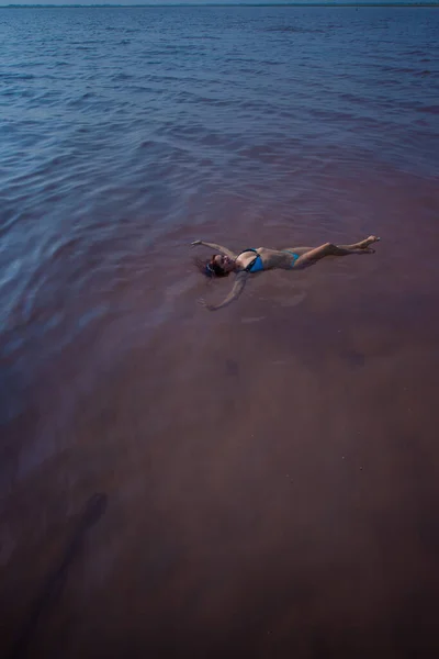 Une femme en bikini bleu nage sur le dos dans un lac salé. Miracle de la nature lac rose. Dépôt et extraction de sel. Eau dense poussant à la surface. Procédures thérapeutiques dans l'eau minérale . — Photo