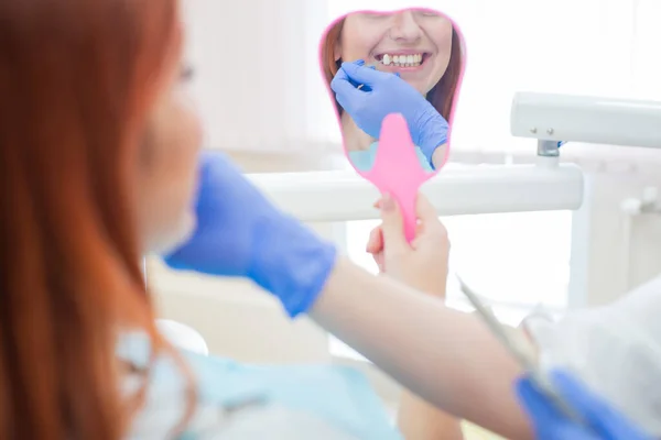 Controllare e selezionare il colore dei denti sulla sedia del dentista. Il dentista fa il processo di trattamento di una bella giovane ragazza dai capelli rossi. Selezione di un impianto dentale.Il riflesso nello specchio . — Foto Stock