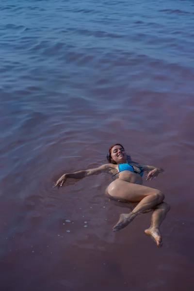 En kvinna i en blå bikini simmar på ryggen i en Saltsjö. Mirakel av naturen rosa sjön. Deponering och extraktion av salt. Tätt vatten som skjuter till ytan. Terapeutiska ingrepp i mineralvatten. — Stockfoto