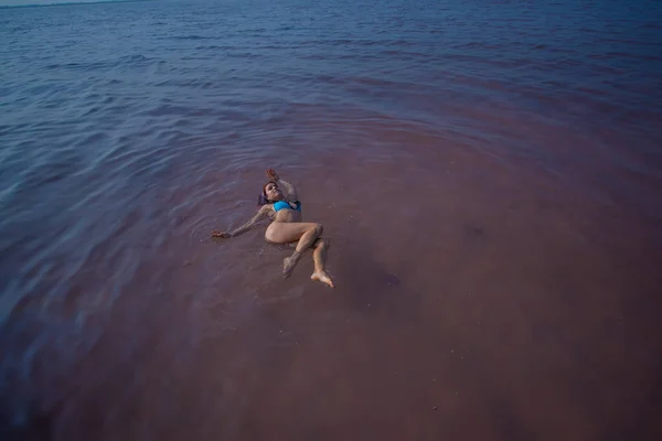 Une femme en bikini bleu nage sur le dos dans un lac salé. Miracle de la nature lac rose. Dépôt et extraction de sel. Eau dense poussant à la surface. Procédures thérapeutiques dans l'eau minérale . — Photo