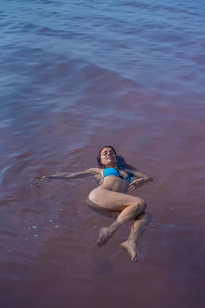 Uma mulher de biquíni azul nada de costas num lago salgado. Milagre da natureza lago rosa. Depósito e extracção de sal. Água densa empurrando para a superfície. Procedimentos terapêuticos em água mineral . — Fotografia de Stock