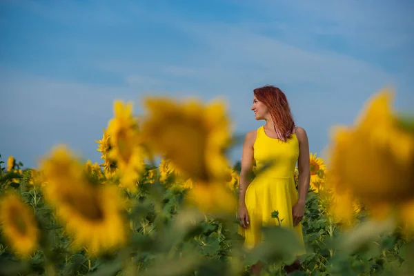 O femeie cu părul roșu într-o rochie galbenă stă într-un câmp de floarea-soarelui. O fată frumoasă într-un soare de fustă se bucură de o zi fără nori în mediul rural. Încuietori roz de păr . — Fotografie, imagine de stoc