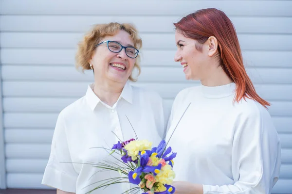 Adult daughter gives flowers to her mother outside, in the courtyard of the house. Spending time together, celebrating at home on weekends. Mothers Day. Warm intergenerational relationships — Stock Photo, Image