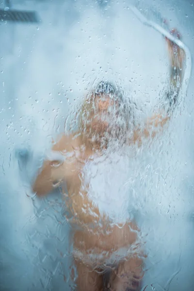 Štíhlá dívka v bílém mokrém tričku se osprchuje a nalije vodu na sklo. Okno s kapkami vody před dívkou. Déšť kapky na okenní sklo s tváří dívky. Sprcha a hygienická lázeňská léčba. — Stock fotografie
