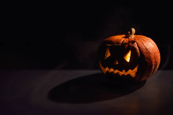 Halloween, oranje pompoen met een eng licht gezicht op een donkere achtergrond. Dikke grijze rook komt naar buiten en verspreidt zich over de zwarte tafel. Een close-up van een zaklamp aan de vooravond van alle heiligen — Stockfoto