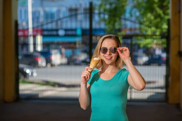 Una mujer feliz en un vestido de color turquesa se para en el patio y se come un cono de gofre en un cálido día de verano. Hermosa rubia en gafas de sol disfruta del helado mientras camina. Postre de verano. —  Fotos de Stock