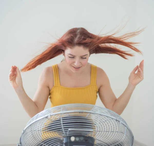 Retrato de una hermosa mujer pelirroja en un mono de mostaza disfrutando de la brisa refrescante de un gran ventilador eléctrico. Una chica sonriente se refresca en el calor caluroso del verano. El cabello se desarrolla por el viento — Foto de Stock