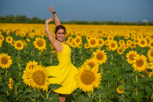 Femeia cu părul roșu într-o rochie galbenă dansând cu mâinile ridicate într-un câmp de floarea-soarelui. O fată frumoasă într-un soare de fustă se bucură de o zi fără nori în mediul rural. Încuietori roz de păr . — Fotografie, imagine de stoc