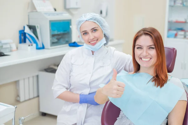 Mulheres felizes um dentista e paciente depois de tratar os dentes no consultório odontológico, sorrindo e olhando para a câmera. Paciente feliz mostra o polegar como um endosso. Equipamento odontológico . — Fotografia de Stock