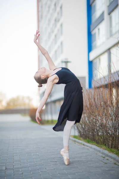 Bailarina em um tutu posando na frente de um edifício residencial de vários andares. Mulher bonita em vestido preto e sapatos pontiagudos com incrível flexibilidade. Linda bailarina executa um elegante — Fotografia de Stock