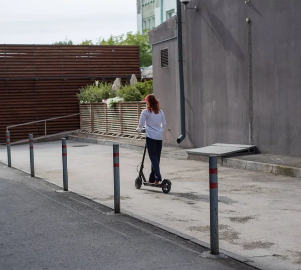 Una ragazza dai capelli rossi con una camicia bianca guida uno scooter elettrico lungo il muro. Una donna d'affari in giacca e cravatta e tacchi rossi gira per la città in un'auto moderna. dress code in ufficio . — Foto Stock