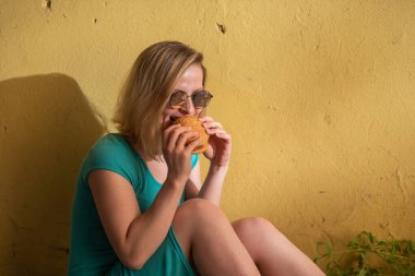 Turkuaz bir elbise içinde mutlu bir kadın kaldırımda oturur ve sıcak bir yaz gününde bir burger yiyor. Güneş gözlüğü güzel sarışın bir konut binanın sarı duvarında önemsiz yiyecek sahiptir.