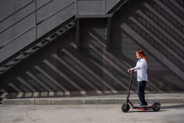 Beyaz gömlekli kızıl saçlı bir kız duvar boyunca elektrikli scooter kullanıyor. Pantolon kostümlü ve kırmızı topuklu ayakkabıgiyen bir iş kadını modern bir arabayla şehirde dolaşıyor. Scooter'da iş kadını.