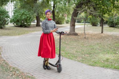 Mor saçlı genç bir kadın parkta elektrikli scooter kullanıyor. Damalı gömlek, uzun kırmızı etek ve papyon lu traşlı bir tapınakla şık kız akıllı telefona mesaj yazıyor. Hipster