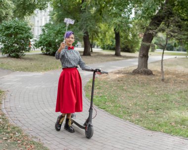 Mor saçlı genç bir kadın parkta elektrikli scooter kullanıyor. Ekose gömlek, uzun kırmızı etek ve papyon bir tıraş tapınak ile şık kız bir selfie sopa kullanarak bir fotoğraf çeker. Hipster.