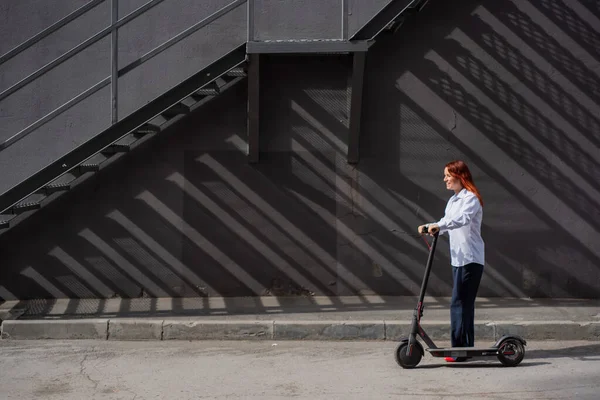 Une fille rousse en chemise blanche conduit un scooter électrique le long du mur. Une femme d'affaires en costume pantalon et talons hauts rouges parcourt la ville dans une voiture moderne. Femme d'affaires sur un scooter . — Photo