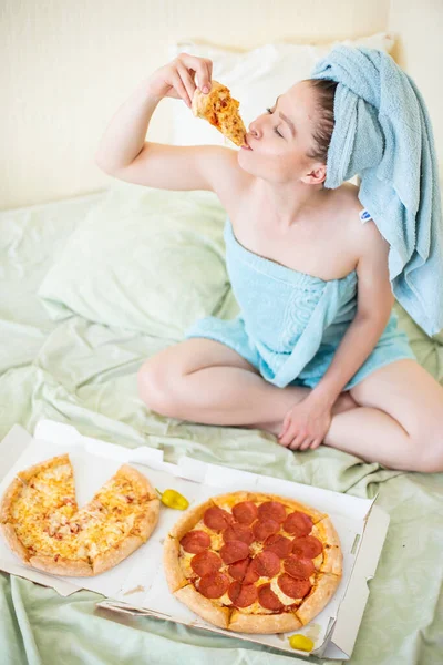 Linda chica con una toalla en la cabeza come pizza en la cama. Mujer joven comiendo comida rápida en la cama. Bodypositive. Mucha comida italiana —  Fotos de Stock