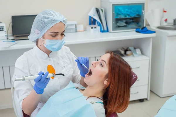 Dentista femenina fija y seca los rellenos dentales con luz. Hermosa dentista femenina poniendo un relleno en un diente con caries de una paciente de cabello rojo —  Fotos de Stock