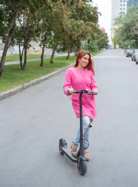 Yırtık kot pantolon ve yüksek topuklu sandalet kırmızı saçlı genç bir kadın yolda bir elektrikli scooter sürmek. Pembe ceketli bir kız hızlı bir şekilde uygun bir elektrikli ulaşım ile şehir etrafında hareket eder. — Stok fotoğraf