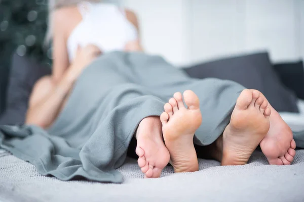 Close-up van mannelijke en vrouwelijke benen. Het liefdevolle koppel ligt en omarmt op het bed onder een grijze deken. Lichte kamer. — Stockfoto