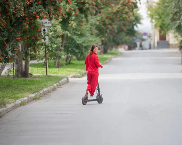Chica pelirroja con un chándal rojo conduce un scooter eléctrico. Una joven con ropa de gran tamaño pasea por la ciudad y escucha música usando auriculares inalámbricos. Vista desde atrás . —  Fotos de Stock