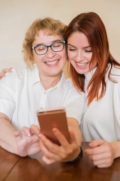 Una anciana madre e hija vestidas con ropa ligera sonríen y se toman selfies. Mamá e hija son fotografiadas en la cámara frontal del teléfono inteligente mientras están sentadas en casa en la mesa . — Foto de Stock