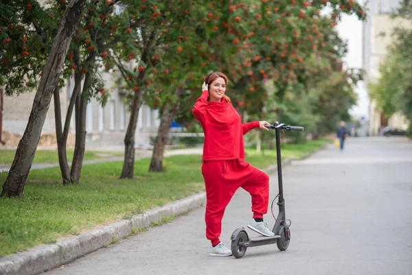 Une fille rousse en survêtement rouge conduit un scooter électrique. Une jeune femme en vêtements surdimensionnés parcourt la ville dans les transports modernes et écoute de la musique à l'aide d'écouteurs sans fil . — Photo