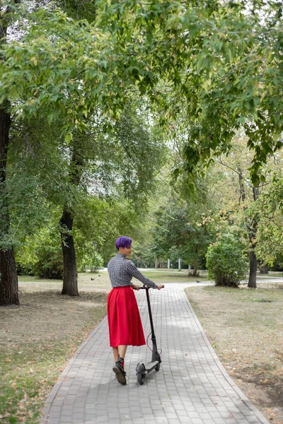 A young woman with purple hair rides an electric scooter in a park. A stylish girl with a shaved temple in a plaid shirt, a long red skirt and a bow tie is riding around the city on a modern device. — Stock Photo, Image