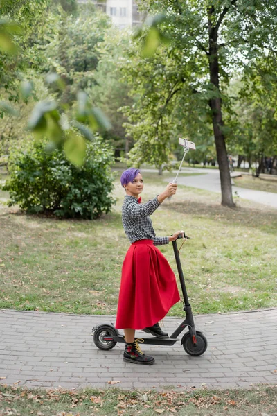 Молода жінка з фіолетовим волоссям їздить на електричному скутері в парку. Стильна дівчина з голим храмом у плетеній сорочці, довга червона спідниця та краватка з бантом робить фотографію, використовуючи паличку селфі. Хіпстер . — стокове фото