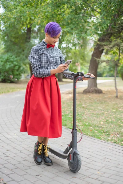 A young woman with purple hair is riding an electric scooter in the park. Stylish girl with a shaven temple in a checkered shirt, a long red skirt and bow-tie writes a message on a smartphone. Hipster — Stock Photo, Image