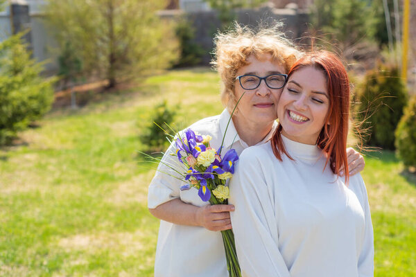 Взрослая дочь дарит цветы своей матери на улице, во дворе дома. Проводим время вместе, празднуем дома по выходным. День Матери. Теплые отношения между поколениями
