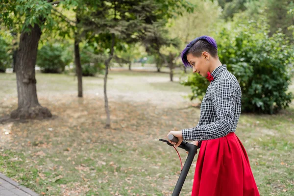 Una joven con el pelo morado monta una scooter eléctrica en un parque. Chica con estilo en una camisa a cuadros, una falda larga de color rojo y una corbata de lazo está montando por la ciudad en un dispositivo moderno. Hipster. . —  Fotos de Stock