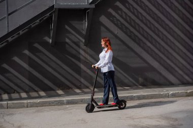Beyaz gömlekli kızıl saçlı bir kız duvar boyunca elektrikli scooter kullanıyor. Pantolon kostümlü ve kırmızı topuklu ayakkabıgiyen bir iş kadını modern bir arabayla şehirde dolaşıyor. Scooter'da iş kadını.