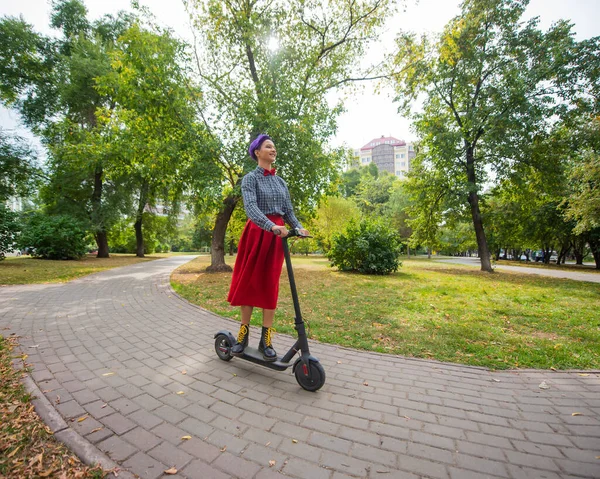 Una joven con el pelo morado monta una scooter eléctrica en un parque. Una chica elegante con un templo afeitado en una camisa a cuadros, una falda roja larga y una corbata de lazo está montando alrededor de la ciudad en un dispositivo moderno . — Foto de Stock