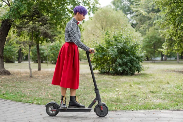 Egy fiatal nő, lila hajú lovagol egy elektromos robogó egy parkban. Egy stílusos lány egy borotvált templom egy kockás ing, egy hosszú vörös szoknya és egy csokornyakkendő a lovaglás a város körül egy modern eszköz. — Stock Fotó