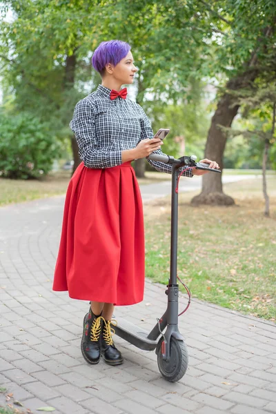 Eine junge Frau mit lila Haaren fährt im Park auf einem Elektroroller. Stilvolles Mädchen mit rasierter Schläfe im karierten Hemd, langem roten Rock und Fliege schreibt eine Nachricht auf ein Smartphone. Hipster — Stockfoto
