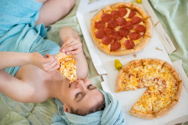 彼女の頭の上にタオルを持つかわいい女の子は、ベッドでピザを食べます。ベッドでピザを食べる若い女性。人生は喜び、体はポジティブです。イタリア料理が大好きです。食習慣、ファーストフード中毒. — ストック写真