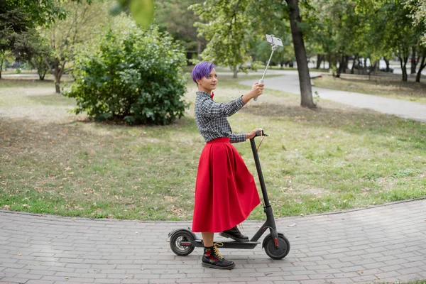 Una joven con el pelo morado monta una scooter eléctrica en un parque. Chica con estilo con un templo afeitado en una camisa a cuadros, una falda larga de color rojo y una corbata de lazo toma una foto con un palo de selfie. Hipster. . — Foto de Stock