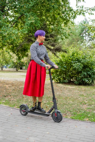 A young woman with purple hair rides an electric scooter in a park. Stylish girl in a plaid shirt, a long red skirt and a bow tie is riding around the city on a modern device. Hipster. — Stock Photo, Image