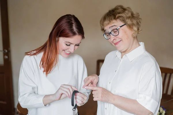 Жінка допомагає старшій матері одягнути фітнес-браслет. Доросла дочка допомагає матері розібратися з пристроєм. Жінка допомагає бабусі налаштувати розумний годинник — стокове фото