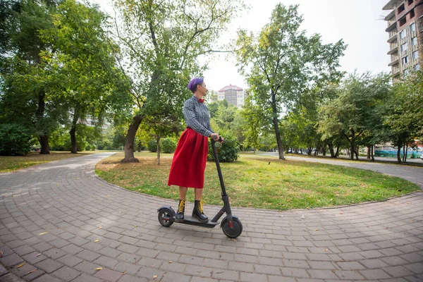 Una joven con el pelo morado monta una scooter eléctrica en un parque. Una chica elegante con un templo afeitado en una camisa a cuadros, una falda roja larga y una corbata de lazo está montando alrededor de la ciudad en un dispositivo moderno . — Foto de Stock